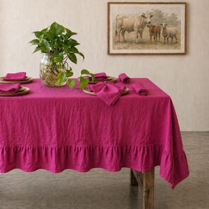 May include: A vibrant fuchsia linen tablecloth with a ruffled edge, set on a rustic wooden table. The table is set with matching napkins and wooden chargers. A framed pastoral painting hangs on the wall, and a glass jar with a green plant sits on the table.
