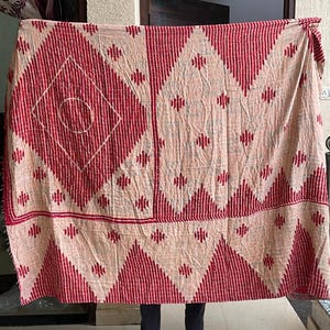 May include: A large, rectangular textile with a repeating geometric pattern. The design features alternating red and beige sections with diamond and zig-zag shapes. The textile is held up, showcasing its full size and intricate details.