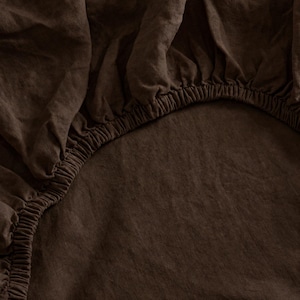 May include: Close-up of a dark brown fitted sheet. The sheet is made of a textured fabric and has an elasticized edge. The fabric is gathered in places, creating soft folds and shadows. The overall look is simple and elegant.