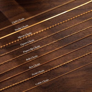 Puede incluir: Una colección de collares dorados exhibidos sobre una superficie de madera. La imagen muestra varios estilos de cadenas, incluyendo cadenas de eslabones, serpiente, espejo, figaro, clip, rolo, caja, trenzada y cable. Cada cadena está etiquetada con su nombre en texto blanco.