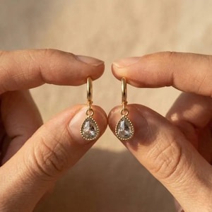 May include: A pair of gold-colored hoop earrings, each featuring a teardrop-shaped, clear gemstone. The gemstones are set within a delicate, beaded frame. The earrings are held up against a neutral background.