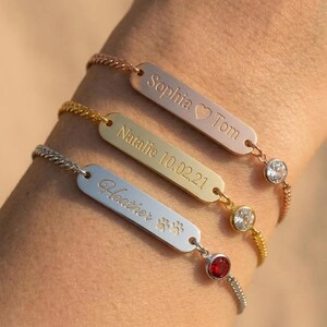 May include: Three personalized bracelets in rose gold, gold, and silver. Each bracelet features a rectangular charm with engraved names and dates. The bracelets have a delicate chain and a gemstone or crystal accent.