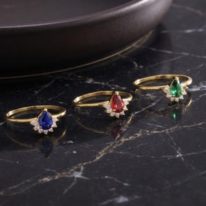 May include: Three gold rings, each featuring a pear-shaped gemstone in blue, red, and green. Each ring is accented with a cluster of small, clear, round-cut stones arranged in a fan-like pattern at the base of the central pear-shaped gemstone.