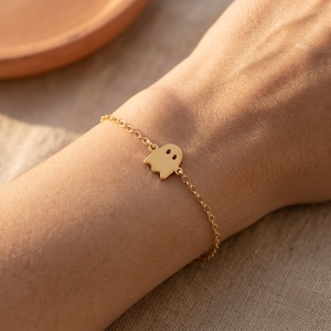 Könnte beinhalten: Ein goldfarbenes Armband mit einem kleinen, flachen, geisterförmigen Anhänger, der mittig an einer zarten, dünnen Metallkette befestigt ist. Der Geisteranhänger hat zwei kleine kreisförmige Aussparungen als Augen und einen gewellten unteren Rand.