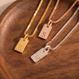 Könnte beinhalten: Drei personalisierte Anhängerhalsketten in Gold, Roségold und Silber. Jeder rechteckige Anhänger hat einen einzelnen Buchstaben: "R", "S" und "W". Die Halsketten werden auf einer Holzoberfläche präsentiert.