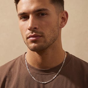 May include: A silver Figaro chain necklace with rectangular nameplates. The necklace is worn by a person wearing a brown t-shirt. The chain has a classic design, with alternating long and short links. The nameplates are engraved with names.