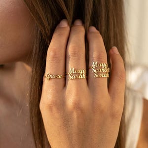 May include: Gold-colored rings with personalized names. The rings are thin and delicate, with the names "Grace", "Maya", "Sarah", "Scarlett" engraved in a cursive font. The rings are worn on the fingers of a hand.