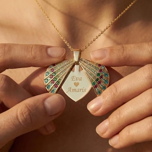 May include: A gold-colored pendant necklace with a heart-shaped locket that opens to reveal the names "Eva" and "Amaris". The pendant has colorful gemstones and is held by a person's hands.