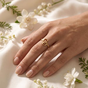 Può includere: Un anello in oro caratterizzato da un design a monogramma stilizzato con le lettere D e P intrecciate. L'anello presenta una finitura metallica lucida e una fascia sottile, indossato su un dito su uno sfondo di tessuto bianco drappeggiato.