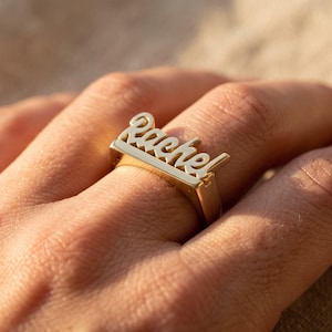 Pode incluir: Um anel de tom dourado com uma base retangular plana rematada com o nome Rachel escrito numa fonte cursiva em relevo. O metal tem um acabamento polido e refletor que capta a luz, criando um design claro e legível.