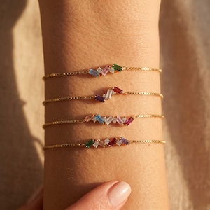Op de afbeelding: Vier gouden armbanden met kleurrijke baguette-geslepen edelstenen en kleine diamantaccenten. De armbanden worden op een pols getoond, waarbij het ontwerp en de kleurvariaties van de sieraden worden benadrukt. De edelstenen omvatten groen, rood, paars en blauw.