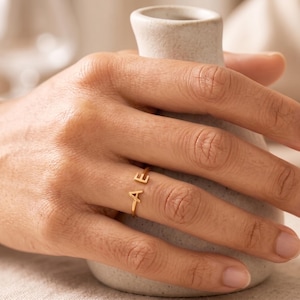 Puede incluir: Un anillo abierto de tono dorado que presenta dos letras mayúsculas, A y E, posicionadas en los extremos de la banda. El anillo tiene un acabado de metal fino y pulido, diseñado para llevarse en el dedo con una estética limpia.