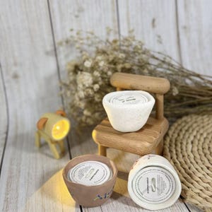Może przedstawiać: Zestaw zapachowych świec w ceramicznych pojemnikach, w tym jedna na miniaturowym drewnianym krześle. Świece mają białe pokrywki z tekstem. Obecna jest również mała, podświetlana lampa w kształcie psa.
