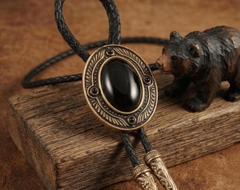 Puede incluir: Una corbata bolo negra con un cordón de cuero trenzado y un colgante ovalado. El colgante tiene un centro de piedra negra y un marco decorativo de color bronce. Una pequeña figura de oso de madera está en el fondo.