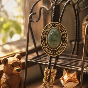 May include: A gold-coloured bolo tie with an oval pendant containing a green stone. The pendant has black accents and hangs from a black braided cord. The tie is displayed on a decorative metal stand.