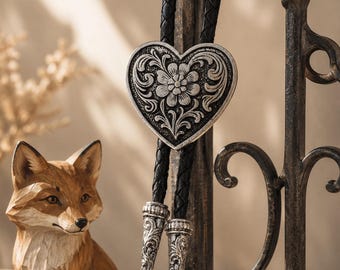 Puede incluir: Una corbata bolo con una pieza central de metal plateado en forma de corazón con un patrón floral en relieve. La pieza está unida a un cordón de cuero trenzado negro que termina en dos puntas de metal plateado con grabados intrincados.