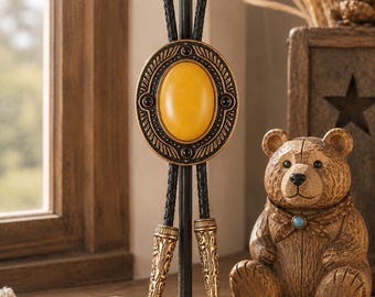 Puede incluir: Una corbata bolo con una piedra amarilla ovalada central engastada en un marco de metal dorado ornamentado con detalles negros. La corbata tiene un cordón de cuero negro trenzado con dos puntas de metal dorado a juego con grabados complejos.