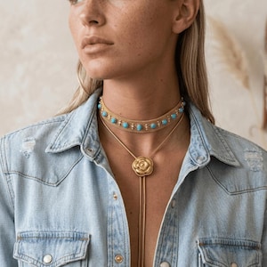 Puede incluir: Conjunto de collares dorados con un choker con piedras turquesas y un collar con colgante en forma de rosa. La joyería se lleva sobre una chaqueta vaquera azul claro.