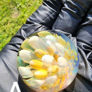 May include: A clear glass paperweight with a floral design. The paperweight features white and yellow petal-shaped elements, creating a layered, three-dimensional effect. The background is a blurred green.
