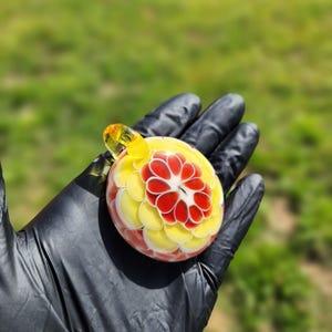 May include: A colorful glass pendant with a floral design. The pendant features a central red flower surrounded by yellow and white petals. It has a yellow loop for a chain. The background is blurred green grass.