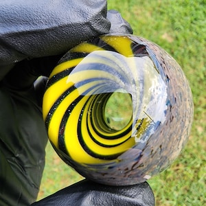 May include: A spherical glass object featuring a hollow center and a swirling pattern of bright yellow and black stripes. The surface is smooth and reflective, showing clear glass sections that create a vortex-like visual effect throughout the piece.