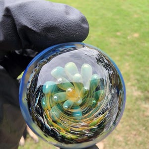 May include: A spherical glass paperweight featuring an internal floral design with layered petals in shades of teal, light green, and yellow. The center contains a small golden orb, surrounded by swirling dark blue and black glass patterns.