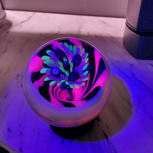 May include: A spherical glass paperweight featuring a swirling internal design with vibrant neon pink and bright green floral-like patterns. The clear glass orb glows under ultraviolet light, highlighting the intricate, multi-colored filaments.