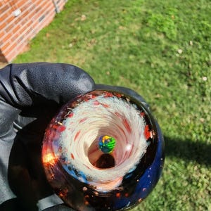 May include: A spherical glass paperweight featuring a deep blue outer layer with embedded red and orange flecks. The center contains a swirling white vortex pattern surrounding a small, iridescent, multi-colored opal-like stone.