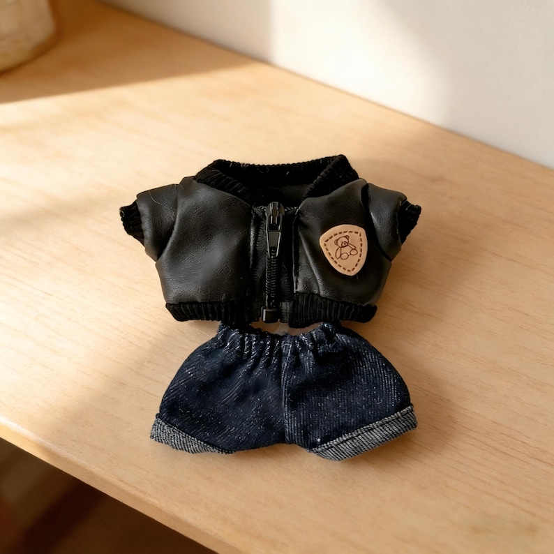 May include: A miniature black faux leather jacket with a zipper and a small patch featuring a bear design. Paired with denim shorts, this outfit is designed for a doll or small plush toy. The jacket has a ribbed collar and cuffs.