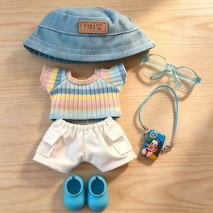 May include: A doll-sized outfit set featuring a light blue bucket hat with "YYDS" text, a striped pastel top, white shorts, blue shoes, glasses, and a phone case with a cartoon character. The set is arranged on a wooden surface.