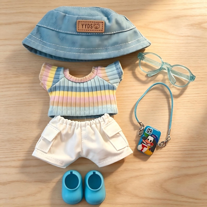 May include: A doll-sized outfit set featuring a light blue bucket hat with "YYDS" text, a striped pastel top, white shorts, blue shoes, glasses, and a phone case with a cartoon character. The set is arranged on a wooden surface.
