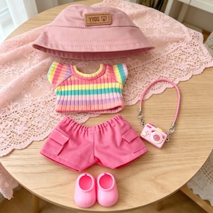 May include: A doll-sized outfit set in shades of pink and rainbow. The set includes a pink bucket hat with "YYDS" text, a striped top, pink shorts, pink shoes, and a camera-shaped purse.