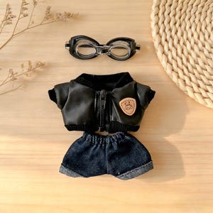 May include: A miniature black leather jacket with a zipper, a denim shorts set, and a pair of black goggles. The jacket has a small patch with a teddy bear design. The items are arranged on a light wooden surface.