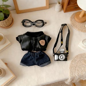 May include: A miniature doll outfit set featuring a black faux leather jacket with a zipper, denim shorts, goggles, and a toy camera with a black strap. The jacket has a small patch. The camera strap says "SMILE".