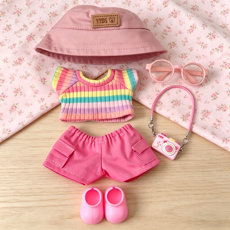 May include: A doll-sized outfit set in shades of pink and rainbow. The set includes a pink bucket hat with "YYDS" text, a striped top, pink shorts, glasses, a camera bag, and shoes. The items are arranged on a floral patterned fabric.