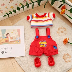 May include: A doll's outfit set comprising a red overall with the text "YOU MADE MY DAY", a striped sweater, and red shoes. The set is displayed on a white rug with a picture of a child's room.