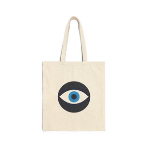 May include: Cream-colored tote bag with a large graphic of an eye. The eye is set within a dark blue circle, with a white iris and a blue pupil. The bag has matching cream-colored handles.