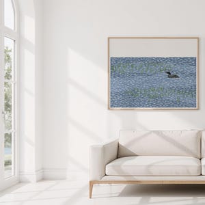May include: A framed wall art print featuring a stylized illustration of a dark-colored loon swimming in blue water with white ripples and patches of green reeds, set against a plain white background within a light wood frame.