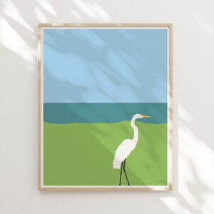 May include: A framed illustration of a white egret standing in a green grassy area with a blue ocean in the background. The sky is light blue.