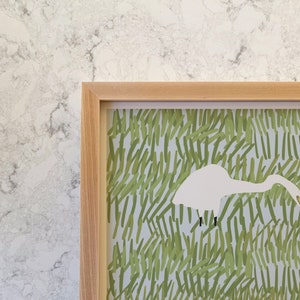May include: A framed print of a white bird with a long beak standing in a field of green grass. The bird is facing the left side of the image and its head is bent down as if it is looking for food.
