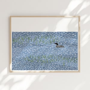 May include: A framed art print featuring a stylized illustration of a black and white loon swimming in a blue lake. The water is depicted with short, horizontal white dashes, and patches of green reeds are scattered throughout the scene.