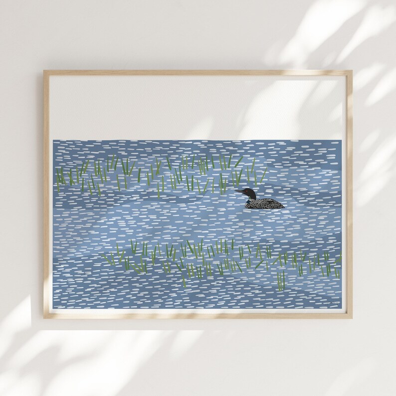 May include: A framed art print featuring a stylized illustration of a black and white loon swimming in a blue lake. The water is depicted with short, horizontal white dashes, and patches of green reeds are scattered throughout the scene.