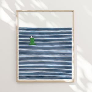 a framed print by artist Jorey Hurley shown hung on a wall with light falling across it. the print is of a seagull perched on a buoy on a background of navy blue and white stripes. The frame is light wood colored.