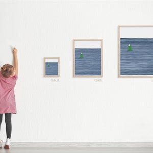 a young girl standing next to three framed art prints by artist jorey hurley. the prints are different sizes . the image on the prints is a coastal scene of a seagull sitting on a buoy with a navy blue striped background.