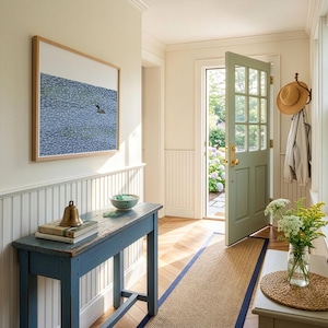 May include: A blue wooden console table holds a brass bell and stacked books. Above, a framed print depicts a bird on blue water. A light green door stands open, while a straw hat and jacket hang on a wall hook nearby.