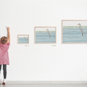 May include: A framed print of a watercolor painting of a heron standing in a body of water with a blue and green striped background. The print is available in three sizes: 6.5 x 12 inches, 18 x 22 inches, and 25 x 31 inches.