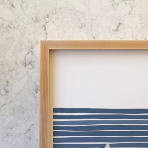 a closeup of the top left corner of a framed print by artist jorey hurley. the frame is solid wood, specifically maple, and is light in color. the visible portion of the art shows nautical navy blue and white stripes and part of a seagull.