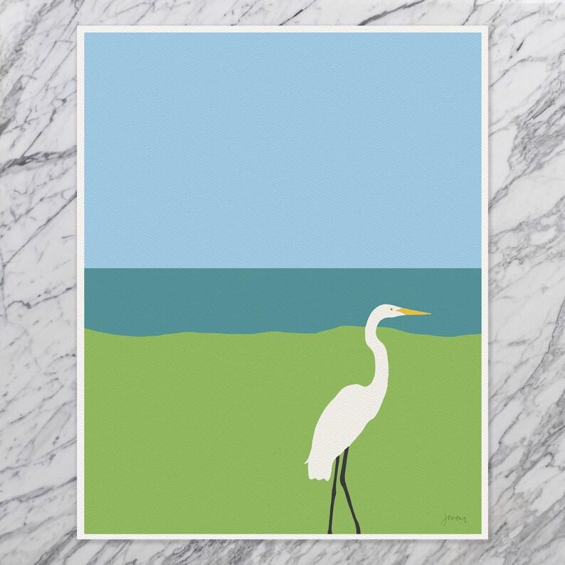 May include: A white egret stands on a green grassy area with a blue ocean and sky in the background. The egret has a yellow beak and black legs.