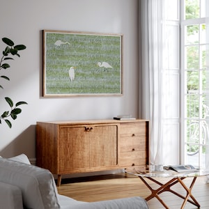 May include: A framed print of a green and white illustration of three white birds in a field of grass. The print is hanging on a white wall above a wooden cabinet with a woven rattan front.