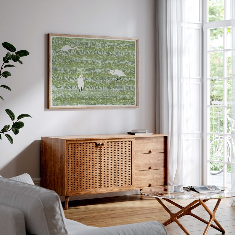 May include: A framed print of a green and white illustration of three white birds in a field of grass. The print is hanging on a white wall above a wooden cabinet with a woven rattan front.